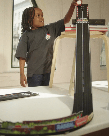 A child plays with a Candylab Roadworks Hairpin Turn kit, racing a toy car down a steep wooden track ramp from a stool.