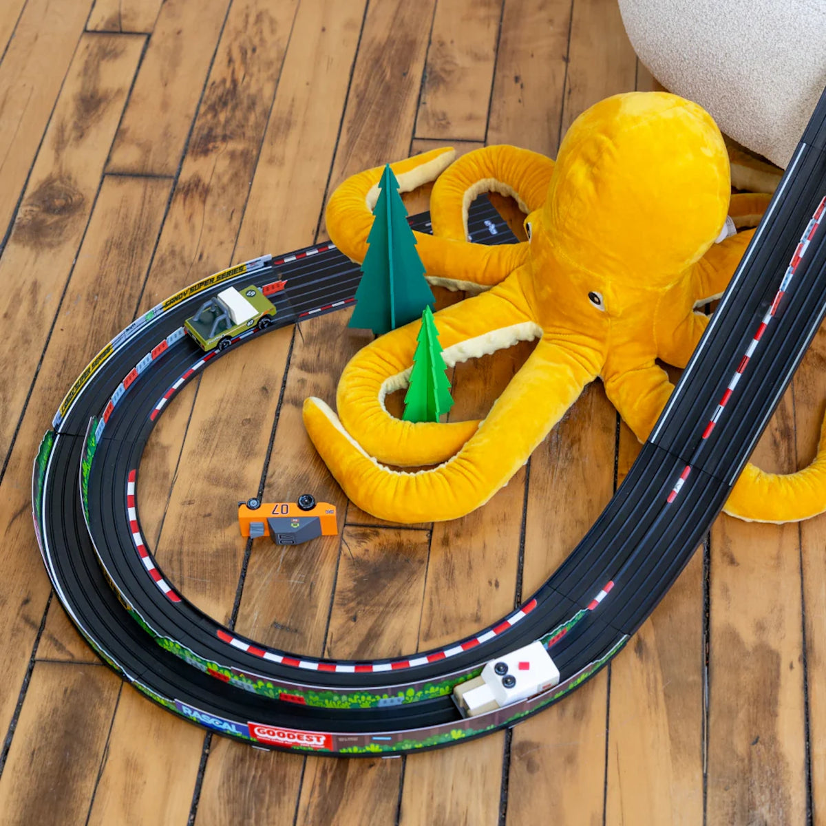 Candylab Roadworks Hairpin Turn race track kit with wooden toy cars, paper trees, and a yellow plush octopus on wood floors.