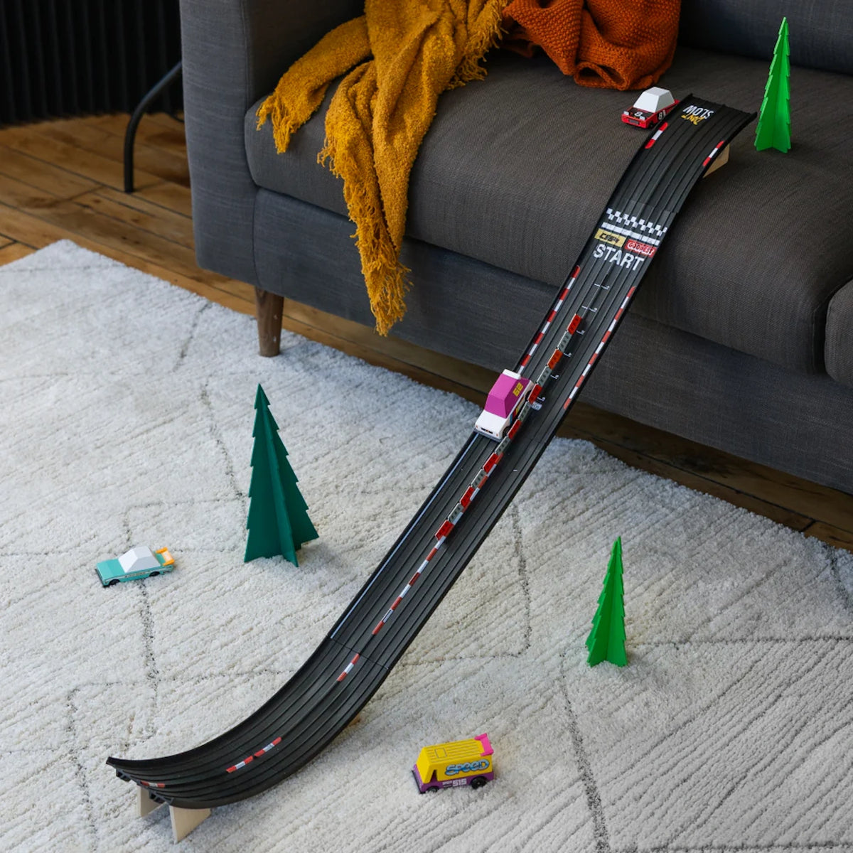 Candylab wooden toy cars racing down a black track ramp leaning on a gray sofa in a modern living room setting.