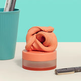 Orange thinking putty twisted into a swirl atop its small container on a desk next to pens.