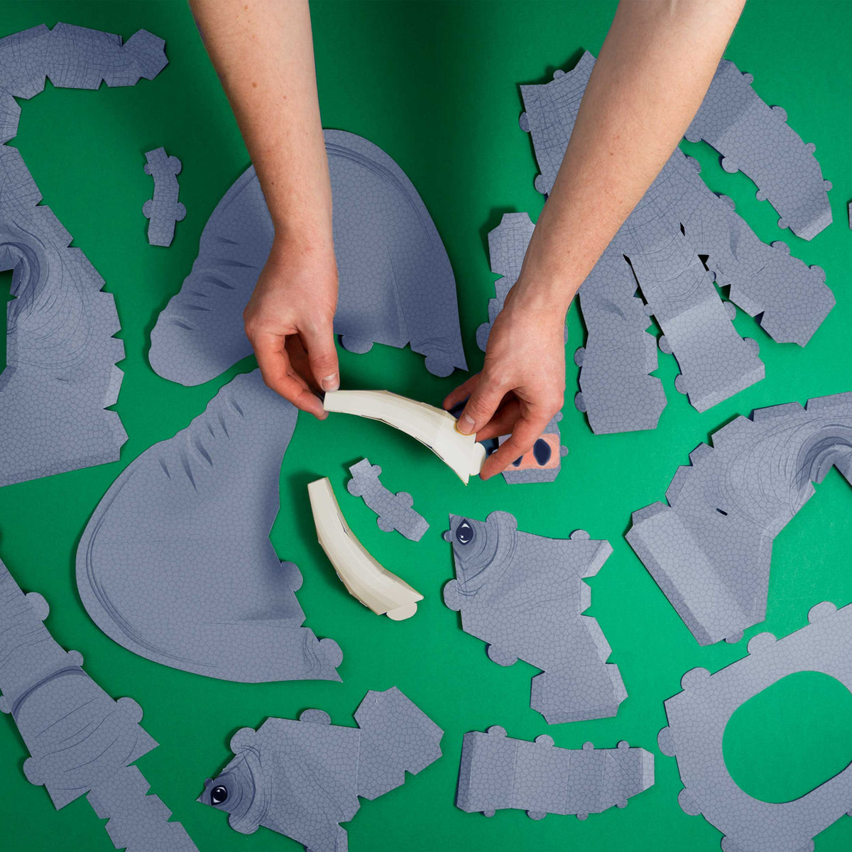 Hands assembling gray, textured cardboard pieces on a green surface. The pieces resemble parts of a 3D model, possibly an animal.
