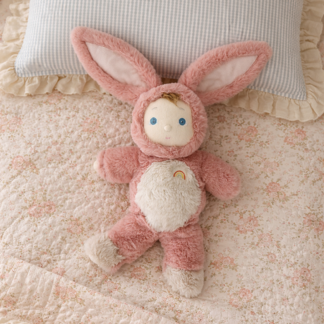 Olli Ella Cozy Dinkum Doll Bunny Pinkie lying on a floral print bed with a blue checkered pillow.
