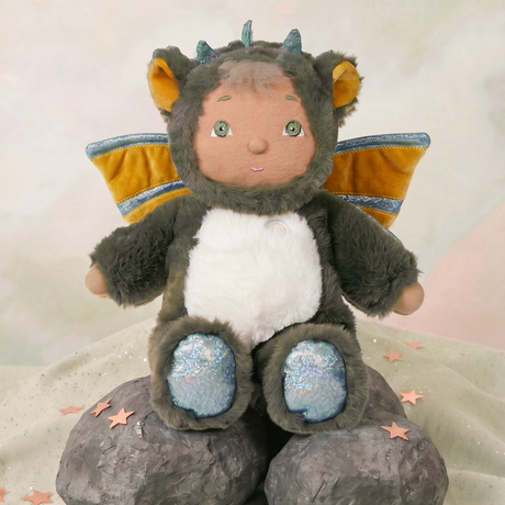 Cozy Day Dinkum Doll Flint wearing a fuzzy green dragon costume with wings, sitting on decorative rocks.
