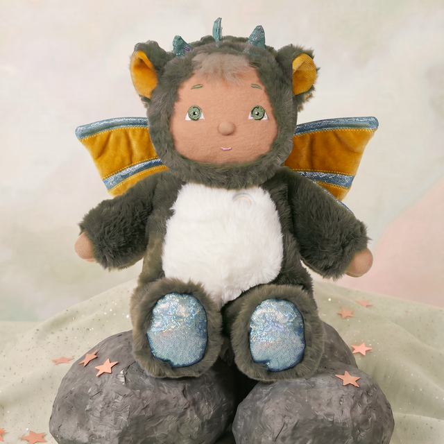 Cozy Day Dinkum Doll Flint wearing a fuzzy green dragon costume with wings, sitting on decorative rocks.