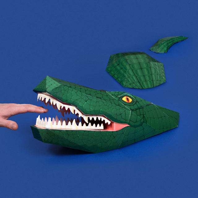 A green papercraft crocodile with its open mouth near a finger, against a solid blue background.