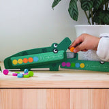 Child playing with a green wooden crocodile color pattern matching game and colorful tokens on a wood surface.