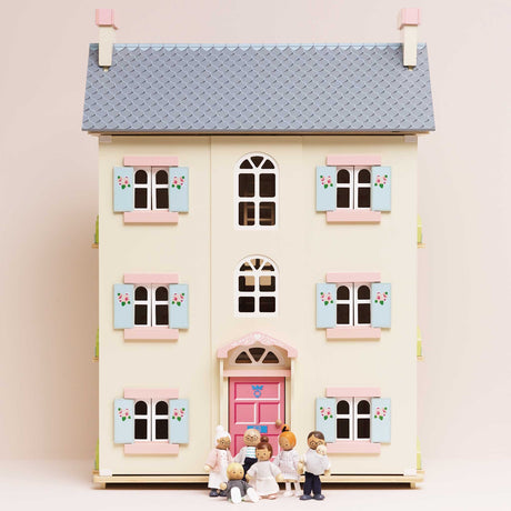 Seven wooden family figures standing in front of a light yellow dollhouse with pink doors and shutters.
