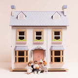 Seven wooden family figures gathered in front of a white dollhouse with a light blue roof.