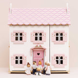 Seven wooden family figures gathered in front of a white dollhouse with pink shutters and a scalloped roof.