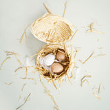 Top-down view of toy wooden eggs in a small woven basket with straw and feathers.