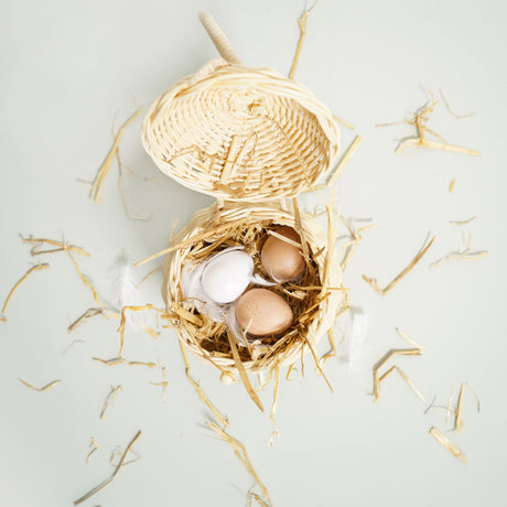 Top-down view of toy wooden eggs in a small woven basket with straw and feathers.