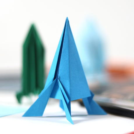 A blue folded origami rocket standing upright with a green paper rocket blurred in the background.