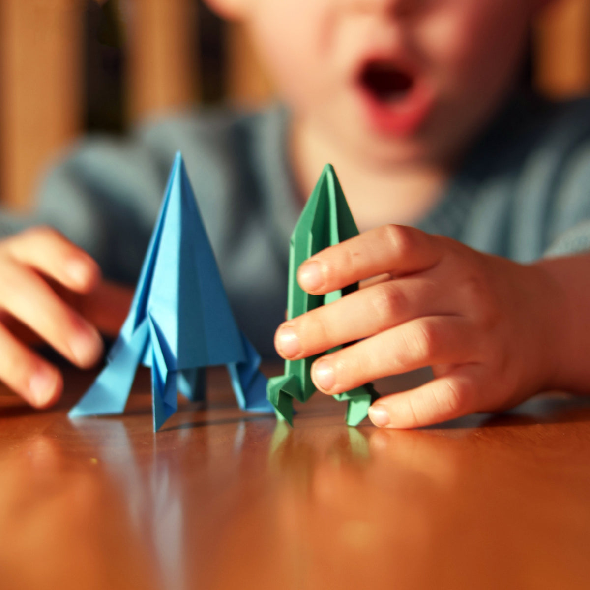 Child's hands playing with blue and green folded paper rockets on a wooden table.