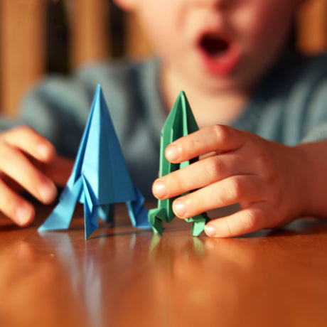 Child's hands playing with blue and green folded paper rockets on a wooden table.