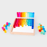 Colorful transparent counters in red, orange, yellow, green, and blue, arranged neatly on a mirrored wooden tray and scattered around it, creating a playful and vibrant tone.