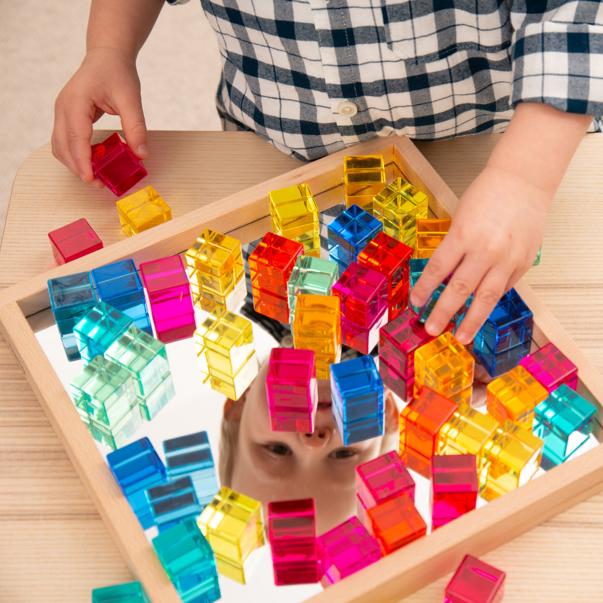 A child in a plaid shirt plays with colorful transparent blocks on a reflective surface, their face partially visible in the mirror, evoking curiosity.