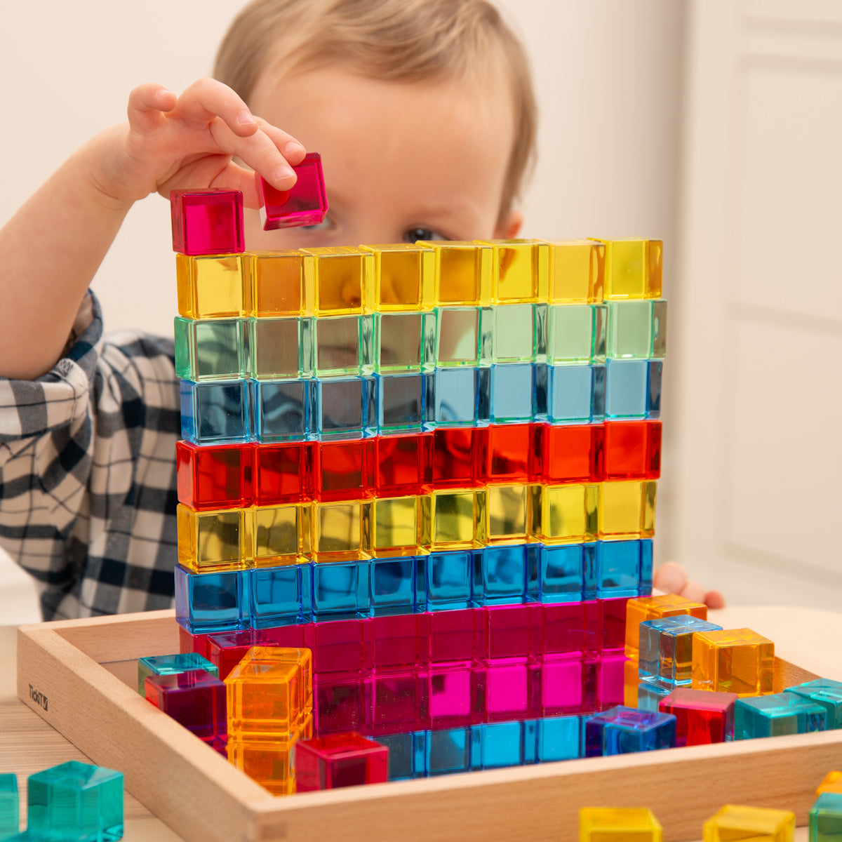 Child building with colorful transparent blocks on a wooden tray. The child is reaching to place a pink block, creating a vibrant, playful scene.