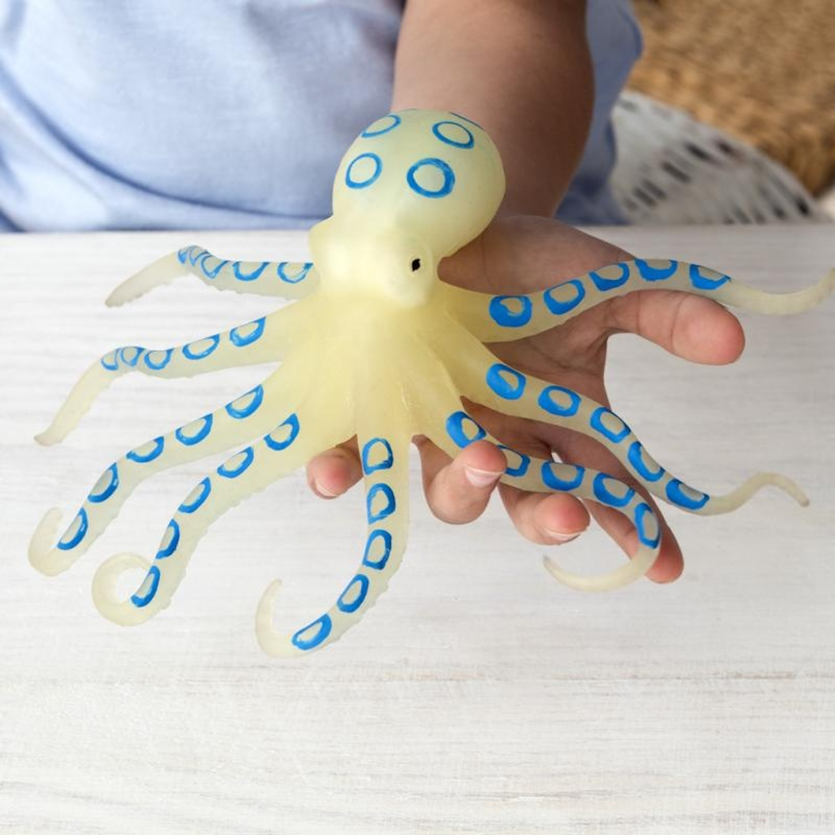A child's hand holds a realistic toy octopus against a light background. The octopus is yellow with blue rings, evoking curiosity and playfulness.