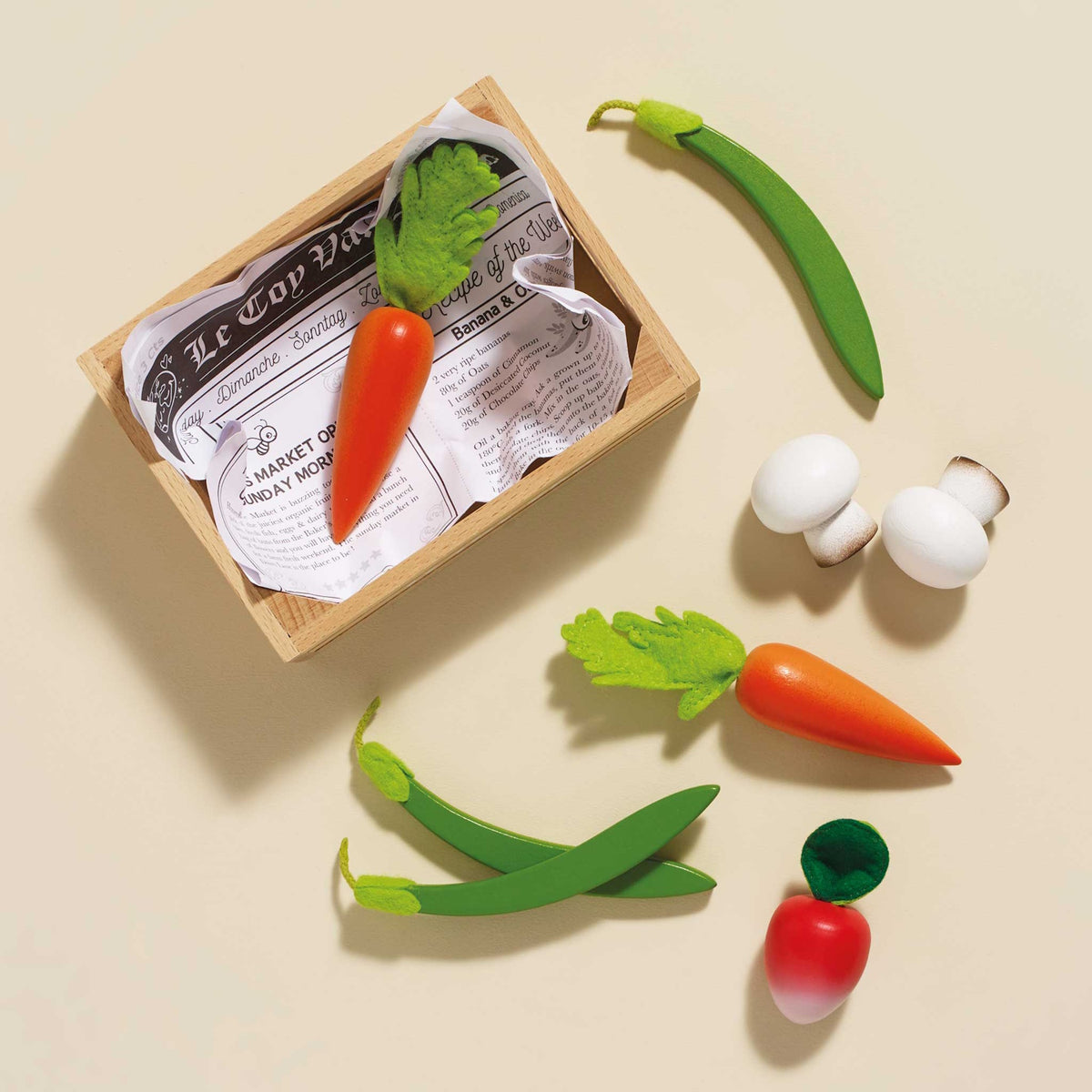 Wooden play vegetables scattered next to a crate lined with play newspaper on a beige surface.