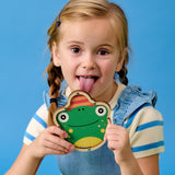 Young girl in blue and white outfit with braids playfully sticks her tongue out while holding a smiling frog cutout, against a bright blue background.
