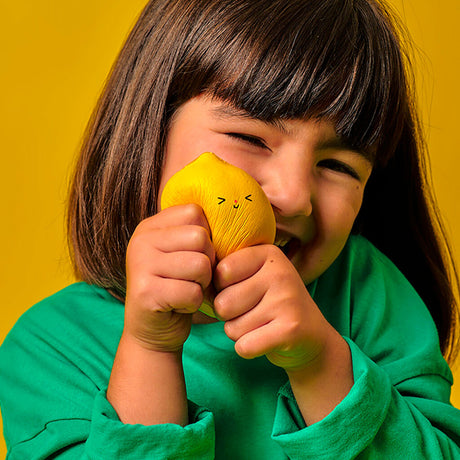 A young child in a green sweater joyfully squeezes a cute, yellow lemon-shaped toy with a smiling face. The vibrant yellow background adds cheeriness.