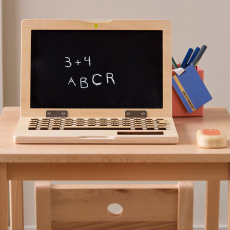 A wooden toy laptop with chalk writing on its screen resting on a child's desk.