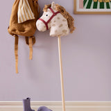 Plush spotted hobby horse toy standing near a wall next to hanging bags and purple boots.