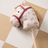 Plush hobby horse with beige spots and a red bridle, resting on a checkered floor.