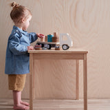 Toddler standing at a wooden table, playing with a wooden toy truck and stacking rings.