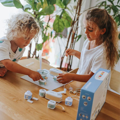 Two young children at a wooden table playing with a modular electronic toy set surrounded by green plants.