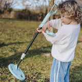 A young child holding and looking at a light green toy metal detector outdoors in a grassy field.