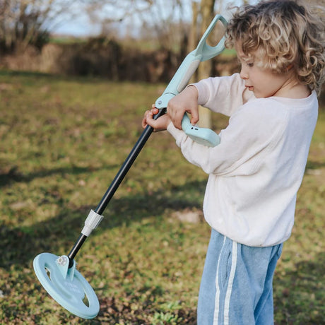 A young child holding and looking at a light green toy metal detector outdoors in a grassy field.