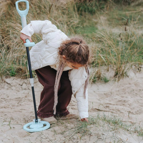 A child using a toy metal detector on a sandy, grassy dune while leaning down to search the ground.