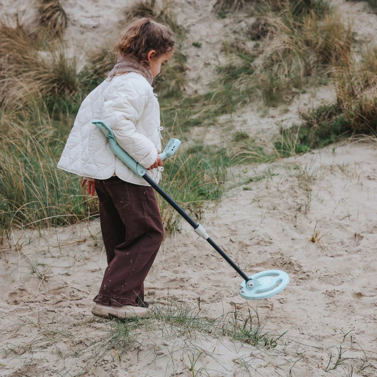 A young girl using a light green toy metal detector to search for treasures on a sandy dune.