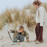 Two children playing with a toy metal detector on a sandy beach by tall grass.