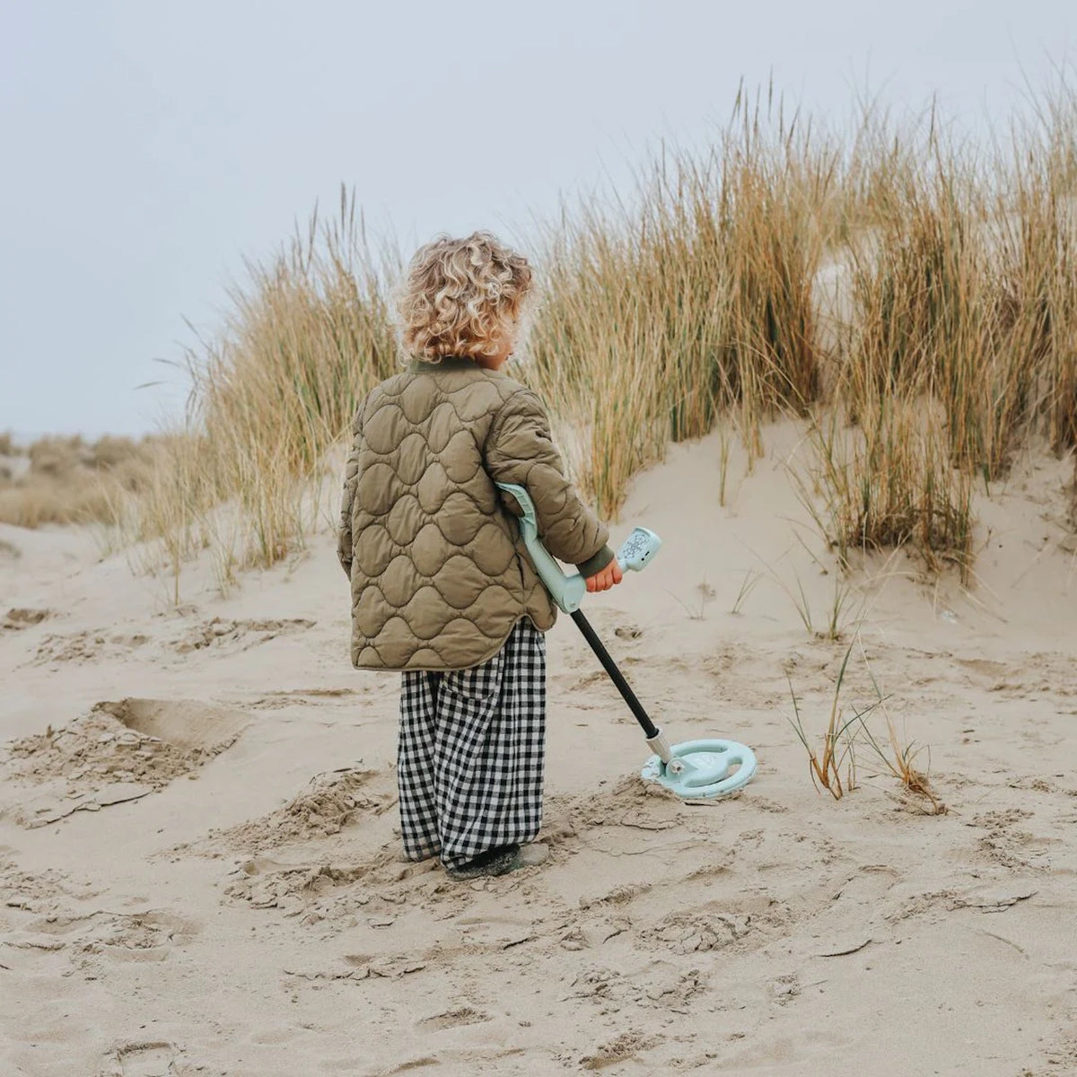 A child from behind wearing an olive jacket, using a light blue toy metal detector on a sandy dune.