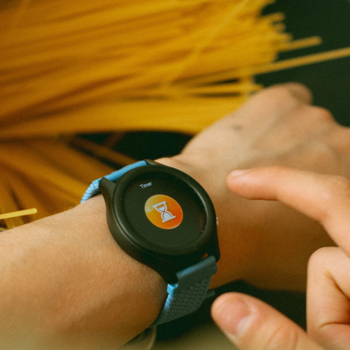 A person sets a timer on a smartwatch, with spaghetti noodles in the background.