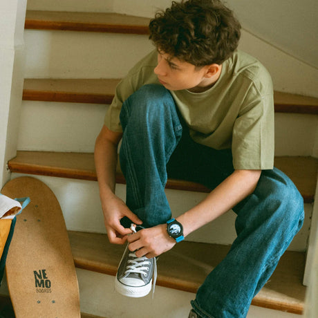 A boy with curly hair sits on stairs, tying his sneaker, with a skateboard leaning nearby.