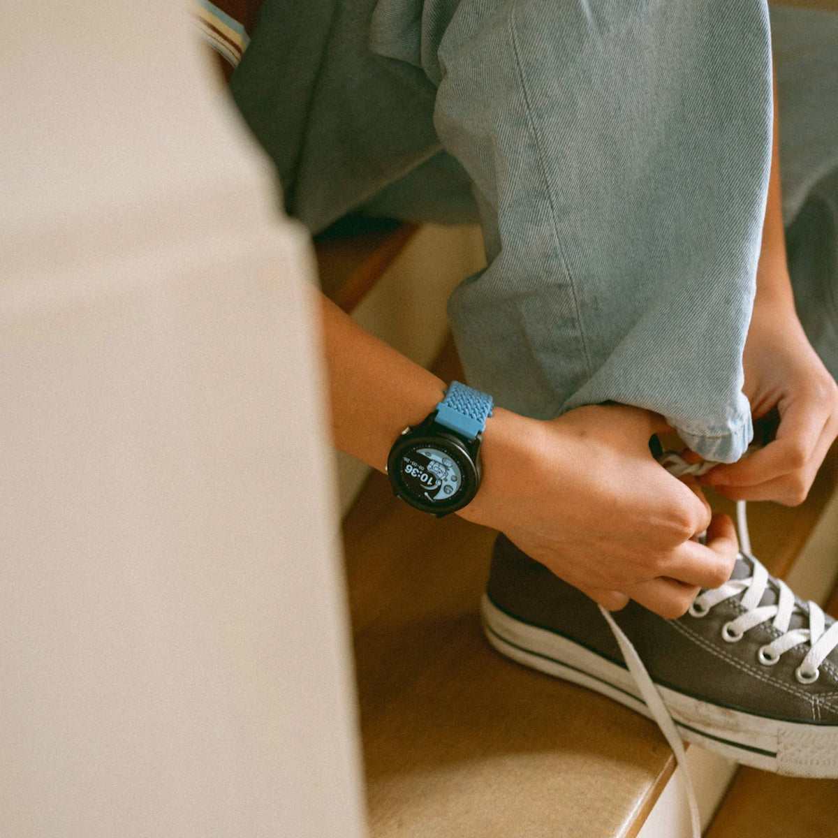 Close-up of a person wearing a smartwatch with a blue strap while tying their canvas shoe on stairs.