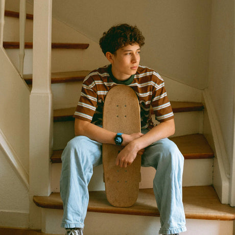 A young person with curly hair sits on indoor stairs, holding a skateboard between their legs.