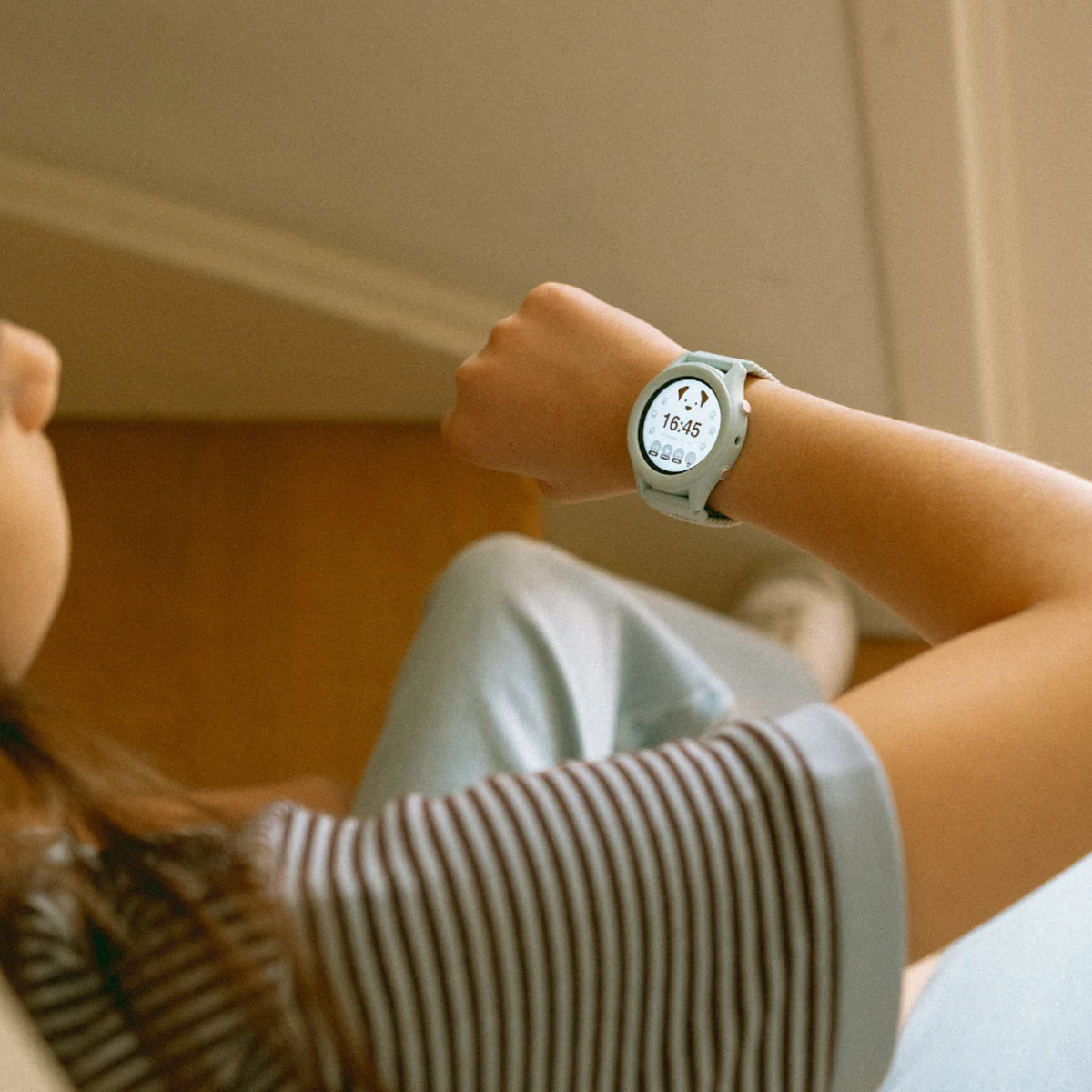 A child looking at a light blue smartwatch on their wrist, showing a dog icon and the time 16:45.