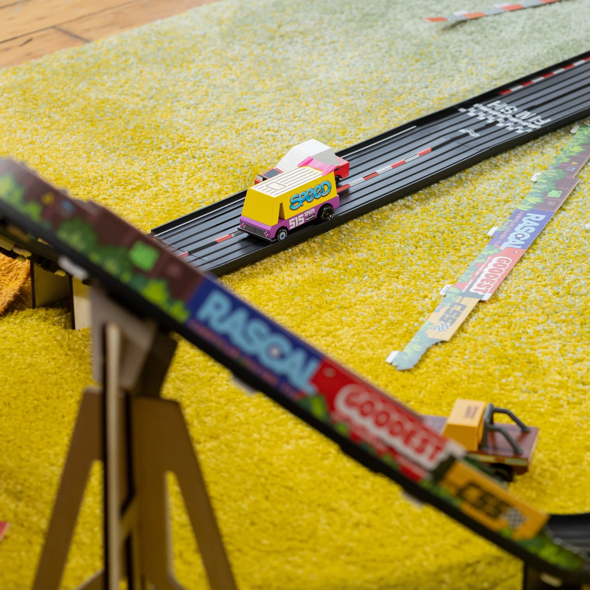 Candylab Roadworks straight track with colorful toy cars racing on a yellow rug, showing a wooden car and track assembly.