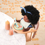 Black baby doll with afro hair in a wooden high chair with toy feeding accessories and play food.