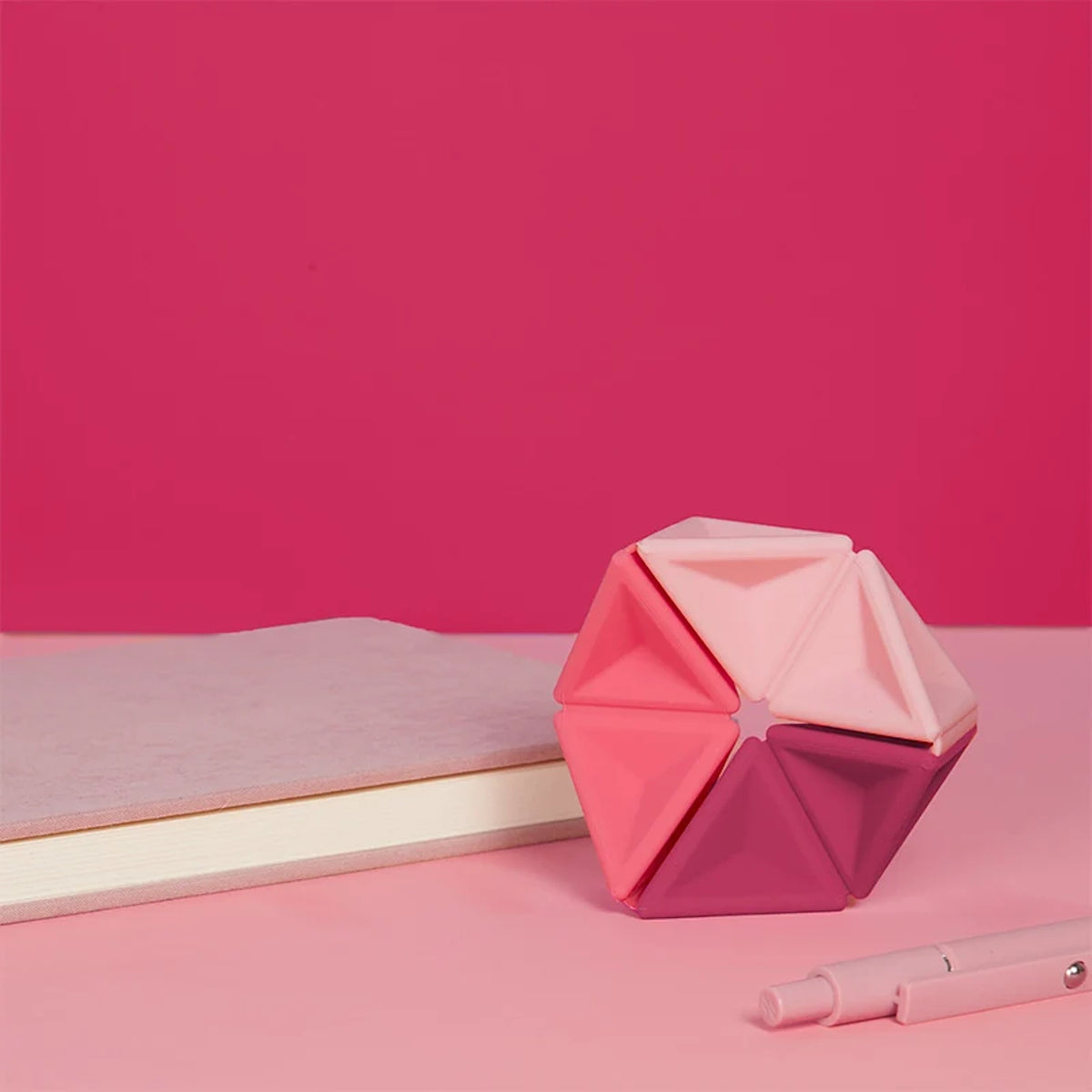 Pink silicone hexaflexagon fidget toy upright on a desk next to a book and pen.