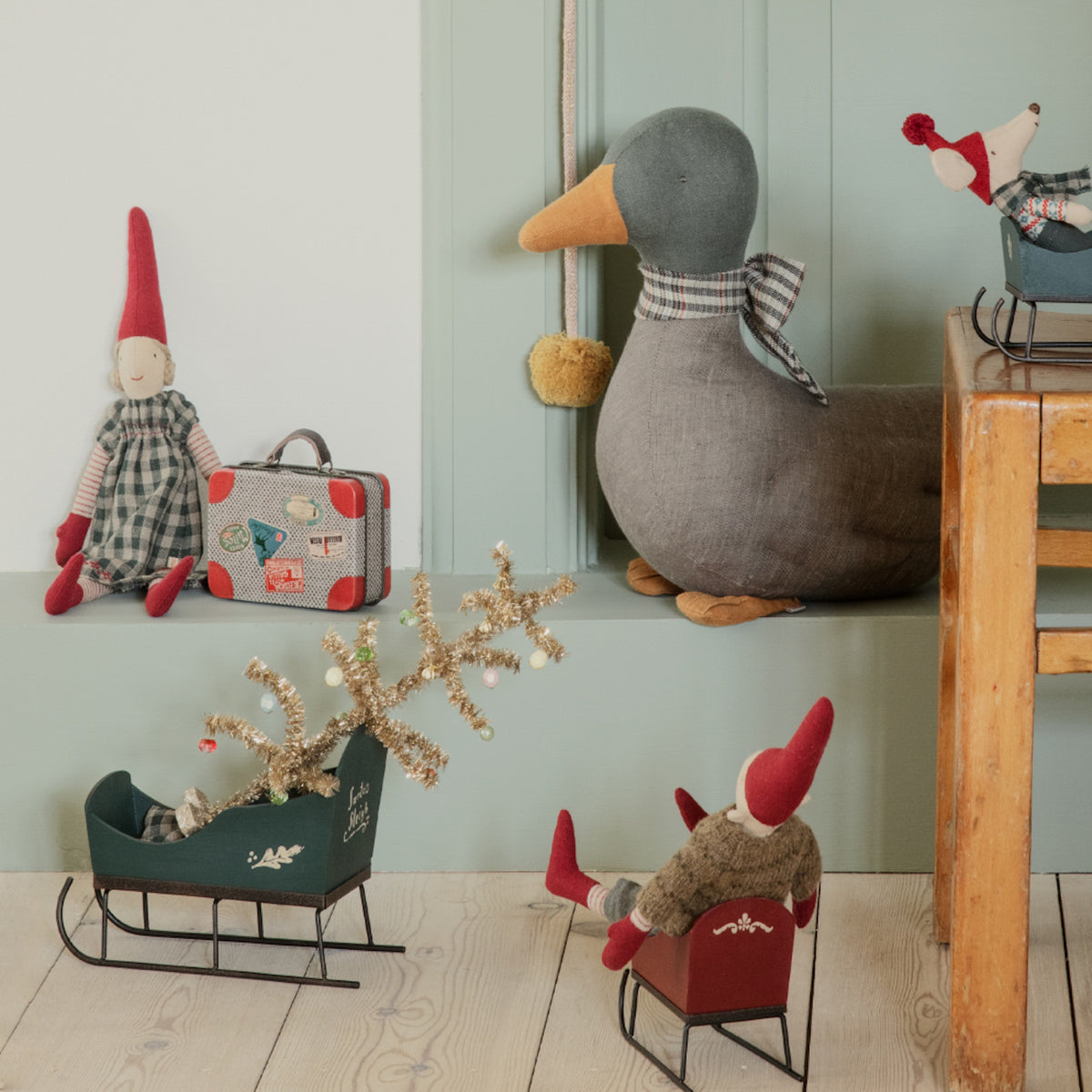 Toy scene with felt gnome, duck, reindeer, and mouse. Gnome holds a suitcase, other toys in sleighs; cozy and whimsical atmosphere.