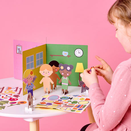 Young girl placing stickers on paper characters playing with a 3D house backdrop on a white table.