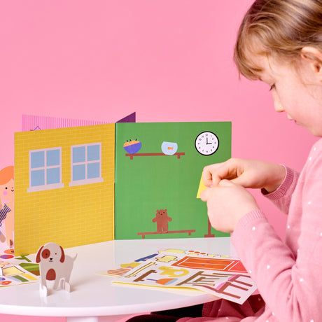 Young girl decorating a colorful 3D paper house playset with stickers on a white table.