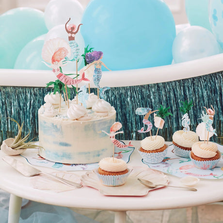 A mermaid-themed birthday cake and cupcakes decorated with toppers against a blue balloon background.