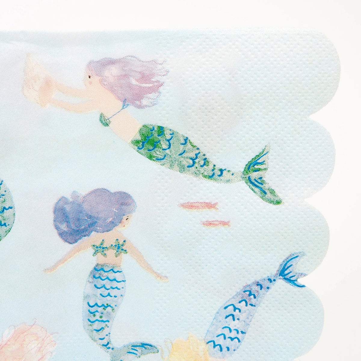 Close-up of a light blue paper napkin with scalloped edges featuring illustrations of swimming mermaids.