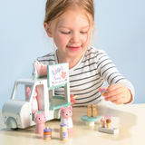 A young girl smiling while arranging tiny wooden food accessories next to a toy milkshake van.